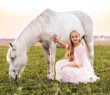 Dziewczynka w różowej sukience siedzi obok białego konia pasącego się na łące