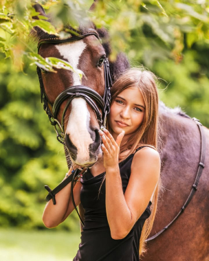 Kobieta w czarnej sukience obejmuje konia na tle zielonych drzew