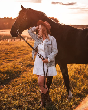 Kobieta w białej sukience, kapeluszu i jeansowej kurtce pozuje z koniem na łące
