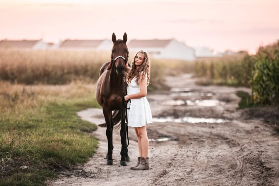 Dziewczyna w białej sukience pozuje z koniem na polnej drodze o zmierzchu