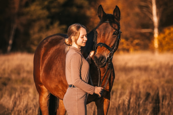 Kobieta w brązowym stroju stoi obok brązowego konia na polu w ciepłym, jesiennym świetle