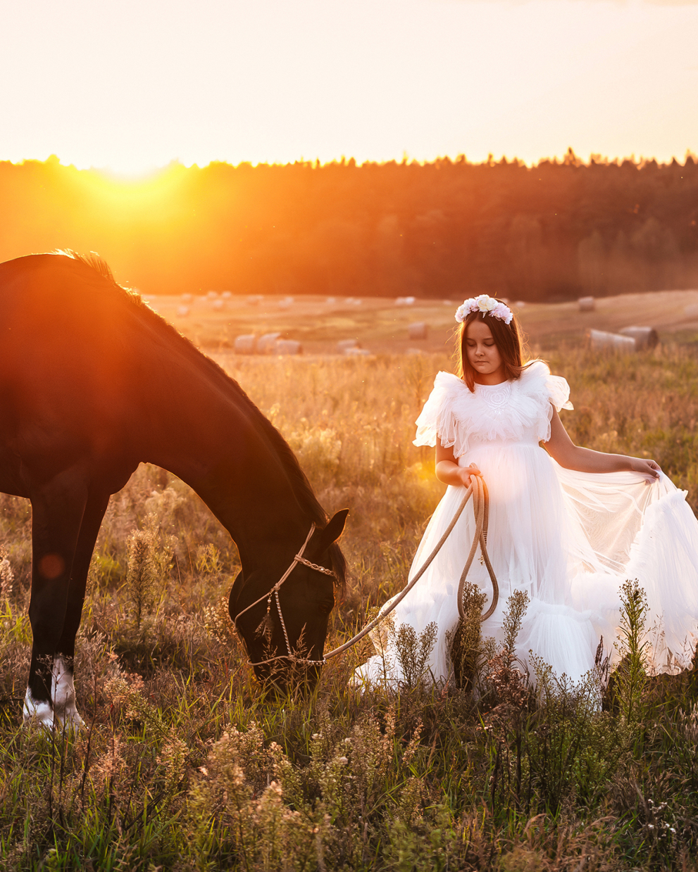 dziewczynka w białej sukni i białym wianku na głowie stoi obok karego konia w halerze boho o zachodzie słońca