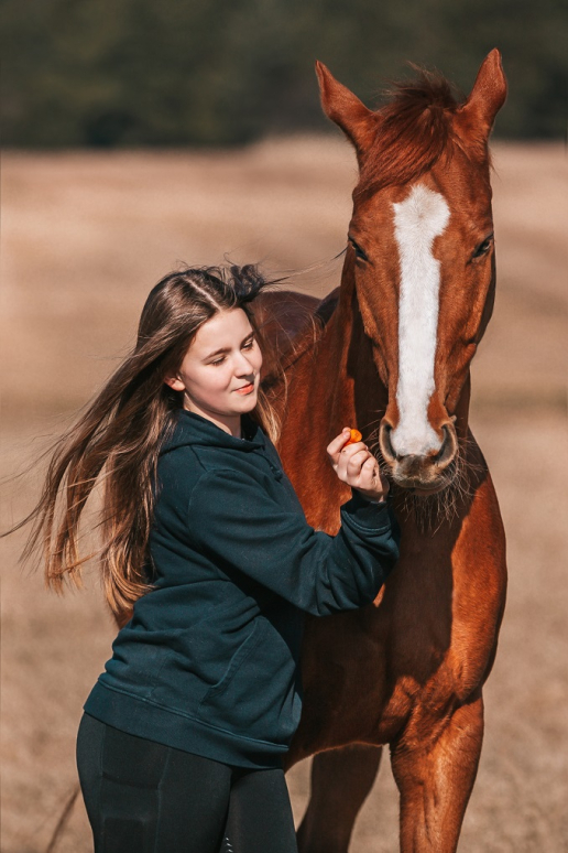 Dziewczyna w czarnej bluzie karmi brązowego konia marchewką na tle pola