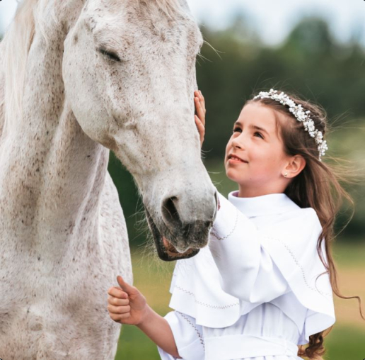 Dziewczynka w białej sukience głaszcze białego konia na tle zielonego krajobrazu