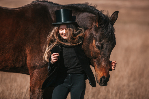 Uśmiechnięta dziewczynka w czarnym ubraniu przytula brązowego konia pasącego się na łące
