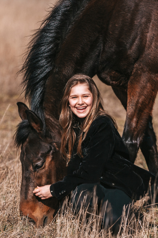 Uśmiechnięta dziewczynka w czarnym ubraniu przytula brązowego konia pasącego się na łące